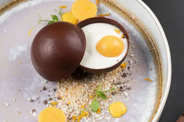 Delicioso trampantojo de comida: una esfera de chocolate oscuro abierta revela un postre cremoso con una yema simulada, rodeado de coco rallado y detalles naranjas.