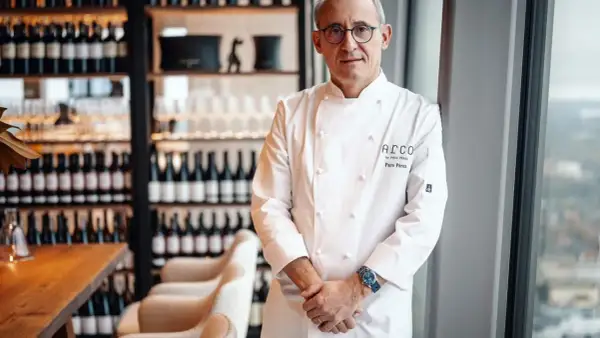 Chef Paco Pérez, con su chaquetilla blanca, posa con confianza en un restaurante elegante, rodeado de botellas de vino.