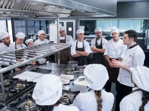 Estudiantes en un curso gestión restaurantes gratis, atentos a las instrucciones de un chef en una cocina profesional.