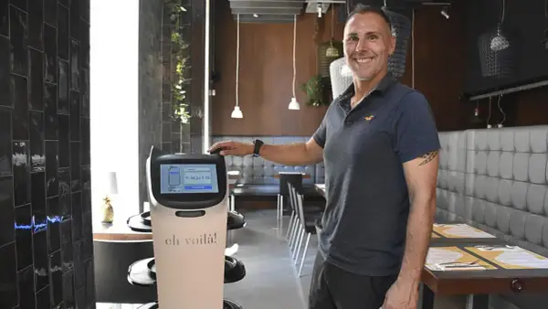Un hombre sonriente junto a un robot que sirve comida en un restaurante moderno.