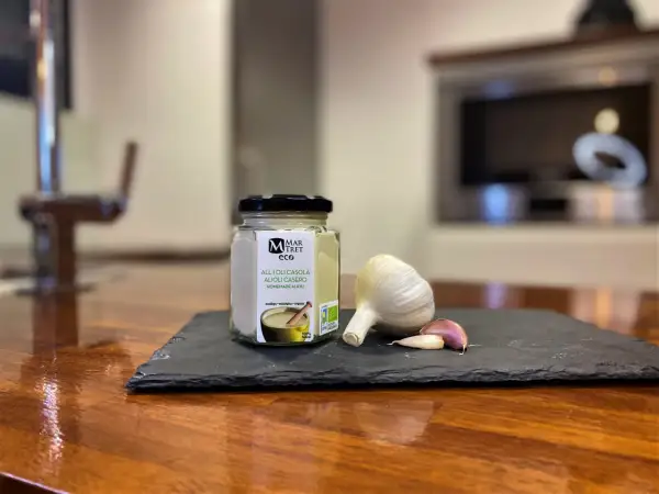 Tarro de alioli casero ecológico junto a una cabeza de ajo y dientes. ¡Este alioli se puede congelar para disfrutarlo más tiempo!