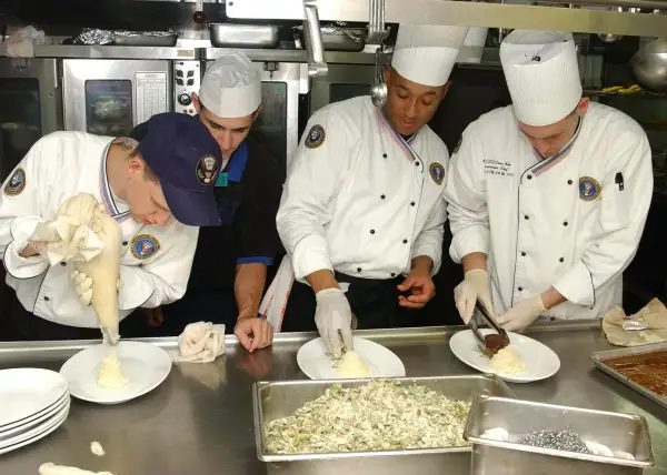 Un ayudante de cocina usa una manga pastelera para decorar un plato. Otros cocineros preparan la comida en una cocina profesional.