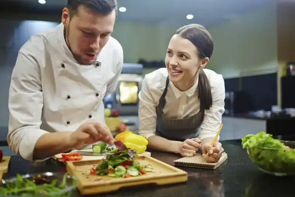 Chef instructor muestra cómo emplatar una ensalada a una alumna atenta en cursos de chef.